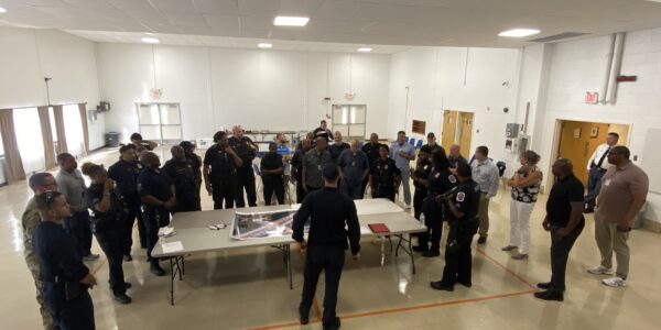 Law enforcement officers and emergency personnel participating in a tabletop exercise for public order management, gathered around a table with maps and plans in a large, well-lit room.