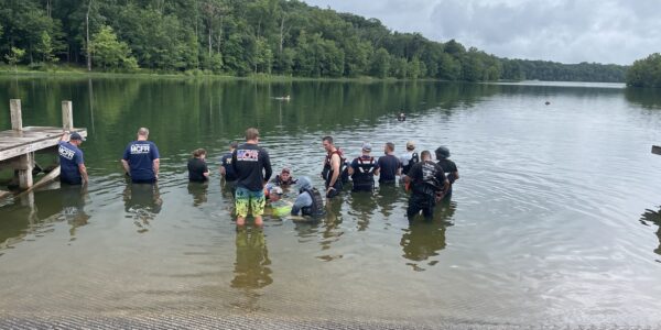 Group of people participating in sonar training at a lakeside, with instructors guiding them in water safety and sonar equipment use, under cloudy skies.